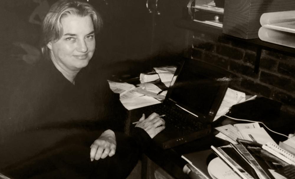 photo of a woman sitting at a cluttered desk, smiling at the camera, with a laptop and papers in front of her